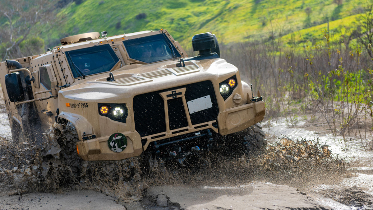 A military vehicle is driving through muddy terrain, splashing water and mud as it moves.