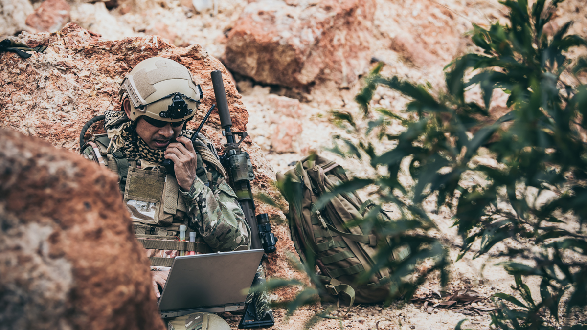 A soldier in camouflage is using a laptop and a radio for communication while sitting amongst rocks in a desert environment.