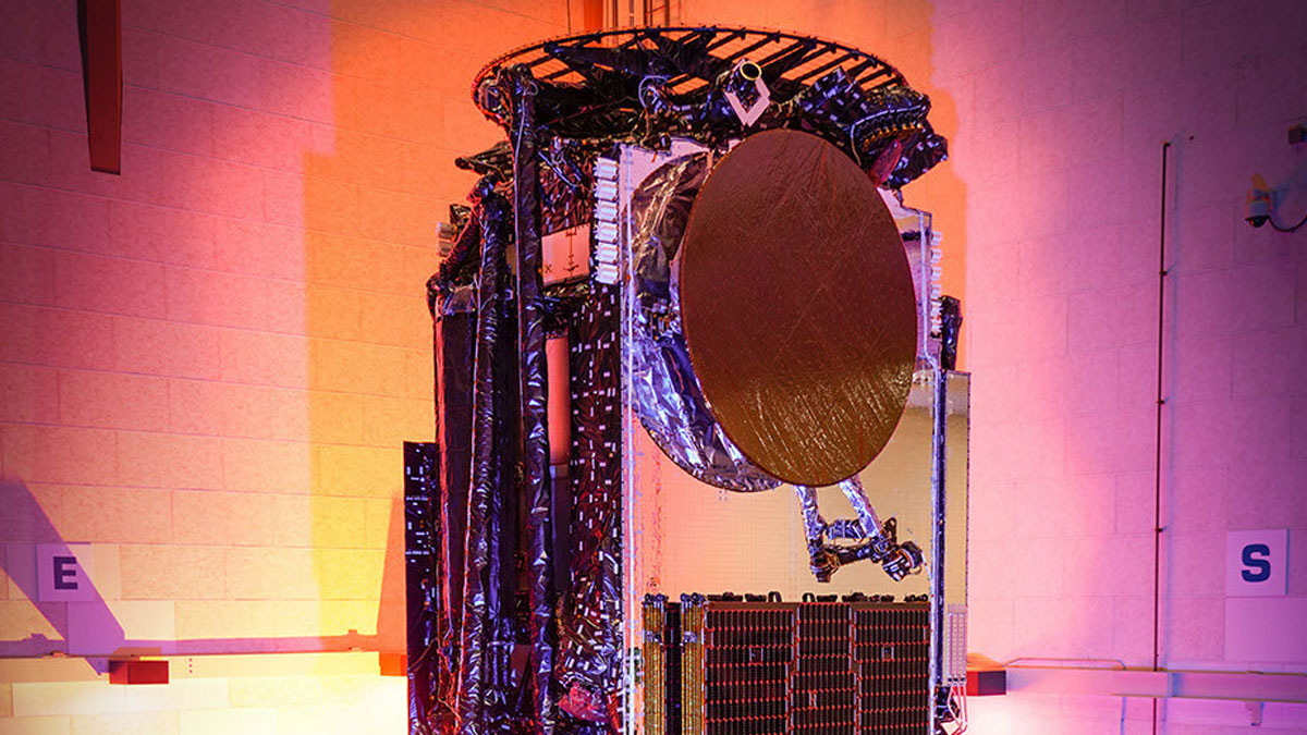 A communications satellite with a large circular dish and multiple panels is subjected to vibration testing in a controlled illuminated environment.