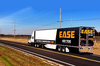 A semi-trailer truck with "EASE" and "KRATOS" branding on the trailer driving on a highway, representing the deployment of self-driving platooning technology by Kratos Defense & Security Solutions.