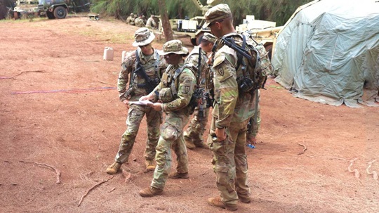 Soldiers in camouflage uniforms review a map together during a training exercise in a wooded area, with military equipment and a tent in the background.