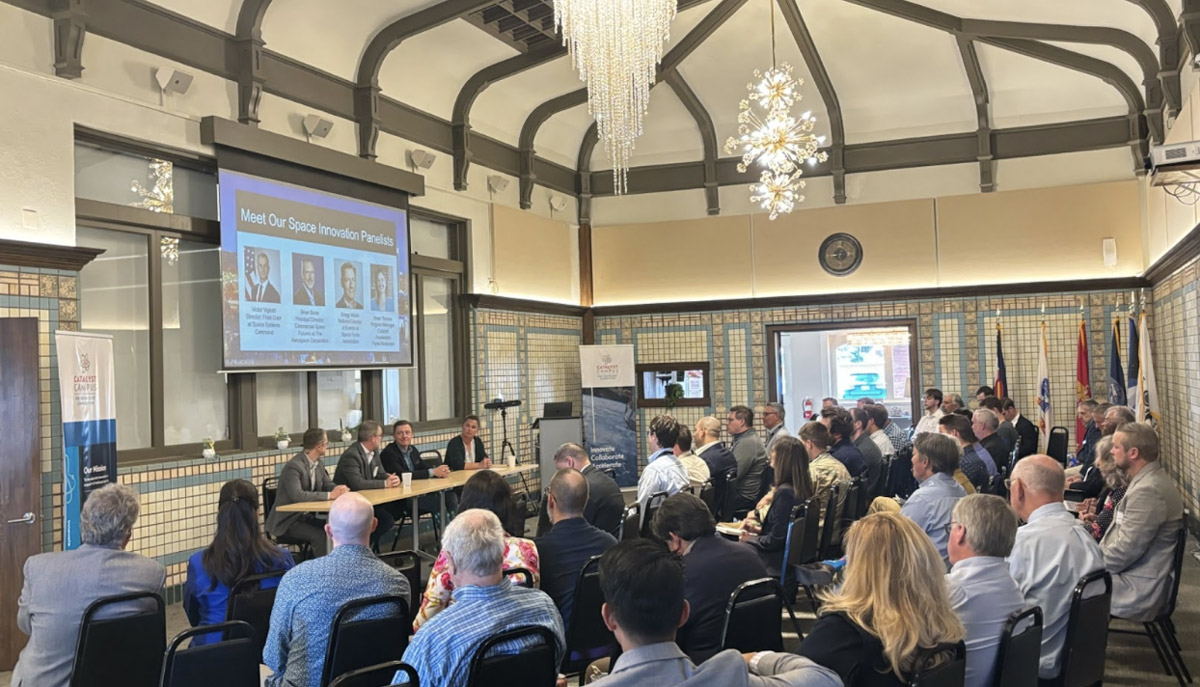 A room full of people attentively listen to a panel of speakers discussing space innovation, with a large screen displaying panelist information and banners indicating the event's association with Catalyst Campus.