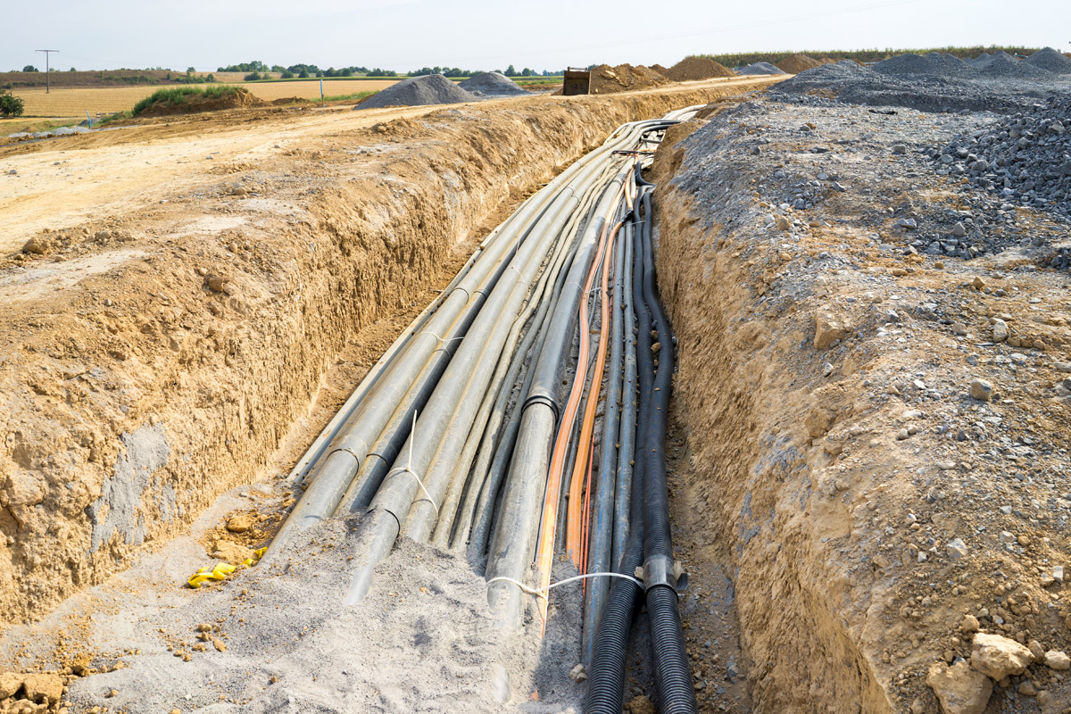 A trench in a rural landscape filled with multiple layers of industrial cables and pipes, showcasing infrastructure development.