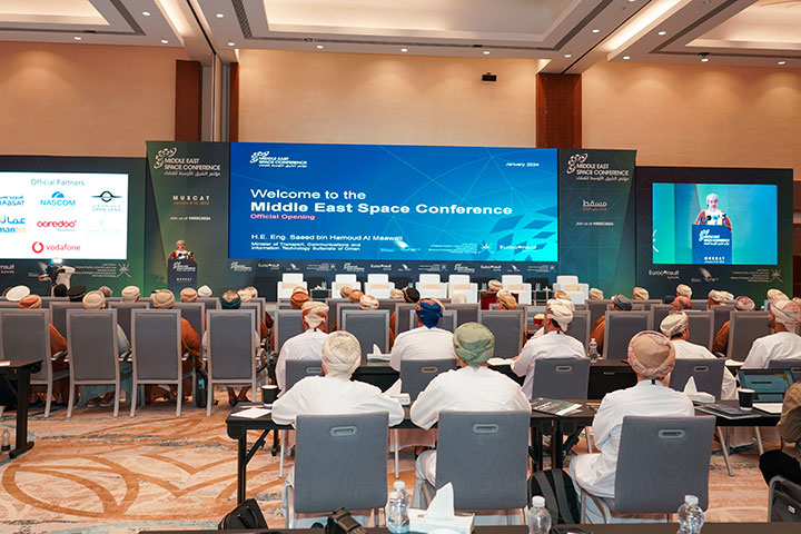 A conference hall with attendees seated and facing a large screen that displays a welcome message for the Middle East Space Conference in Muscat. The stage is adorned with banners featuring the logos of various sponsors and partners.