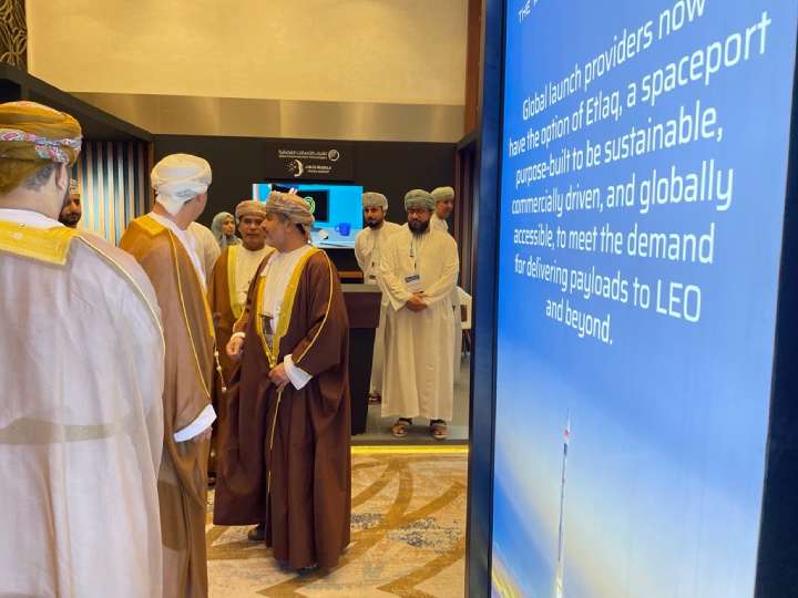 Government representatives engaged in conversation at the Middle East Space Conference in Muscat, Oman, with a presentation in the background discussing a spaceport designed for sustainability and global accessibility.