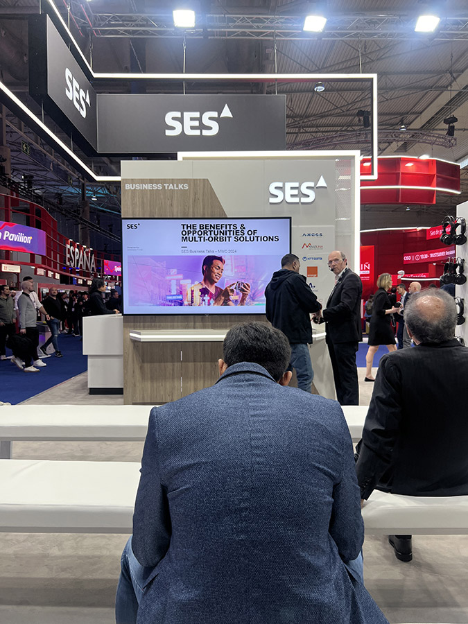 Attendees are seated and standing around a presentation area with a screen displaying 'THE BENEFITS OF MULTI-ORBIT SOLUTIONS' at the SES exhibit.