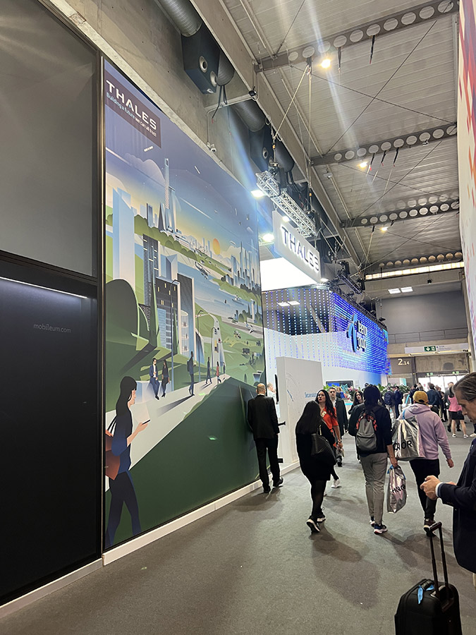 A bustling exhibition hall with people walking past a large Thales advertisement featuring a futuristic cityscape.