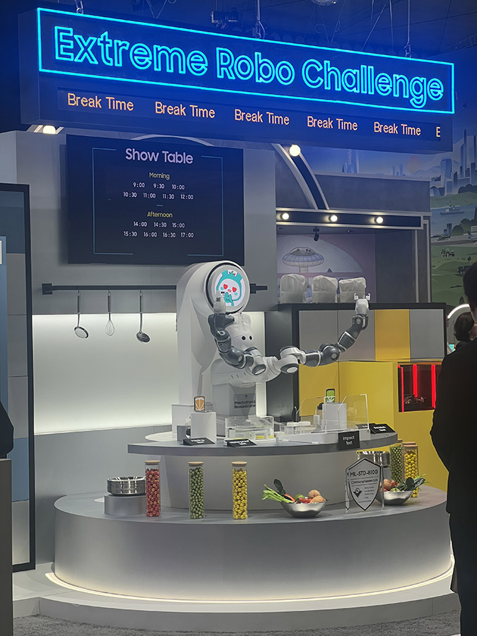 A robot arm performing a demonstration at the 'Extreme Robo Challenge' booth with a schedule displayed above.