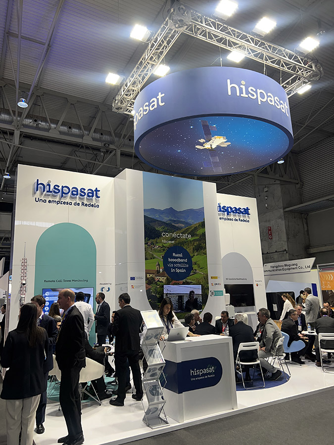 People engaged in conversation at the Hispasat booth, which features branding and images of satellites and nature.