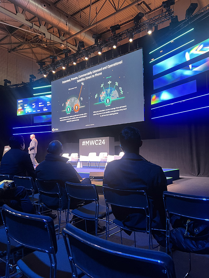 A presenter speaking at a conference with a slide titled 'Potential Power, Commercially Licensed and Operational' showcasing satellite to phone technology.