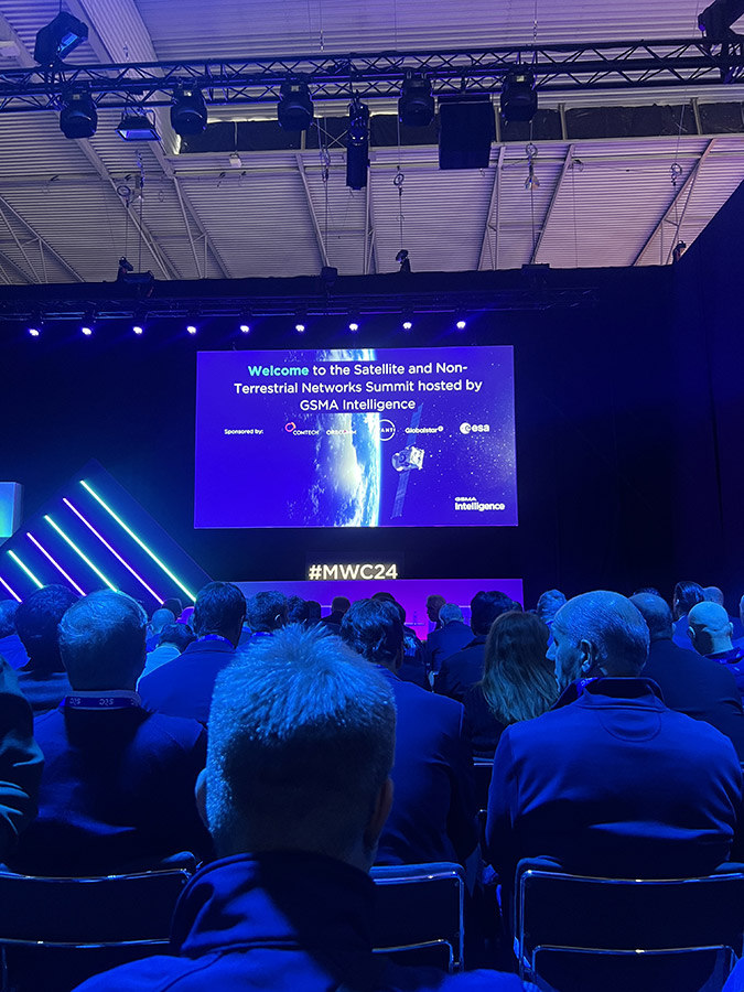 A crowd watching a presentation with a welcoming slide for the 'Satellite and Non-Terrestrial Networks Summit' hosted by GSMA Intelligence.