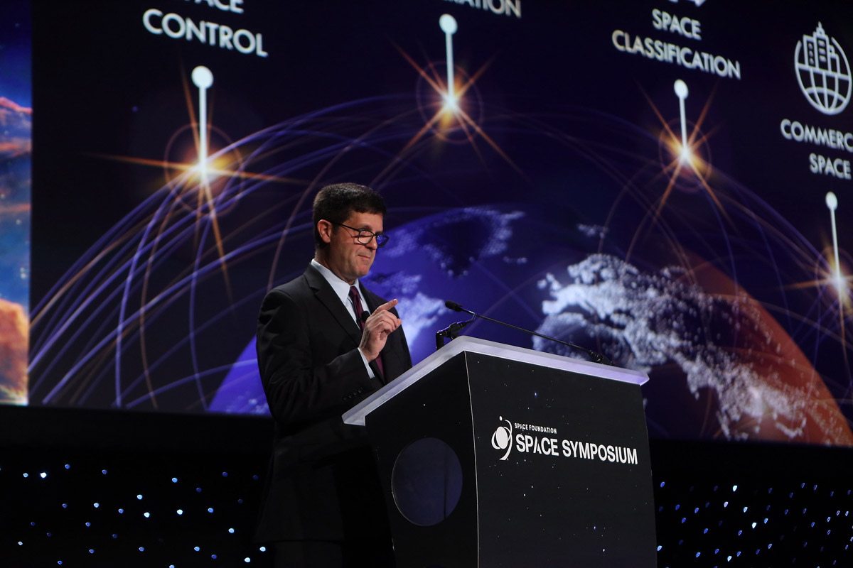 A man is speaking at a podium labeled 'Space Symposium' with a backdrop displaying space-related themes and text, such as 'Space Control' and 'Space Classification'.
