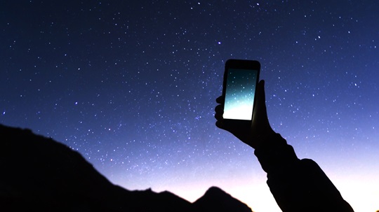 A hand holding up a smartphone displaying an image of the Milky Way against a real starry night sky in the background.