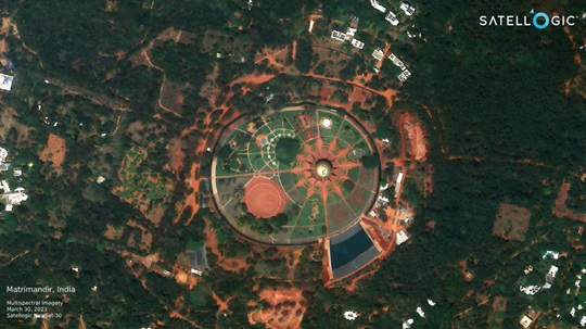 A high-resolution satellite image of the Matrimandir temple in Auroville, India, showing its circular structure surrounded by gardens and greenery.