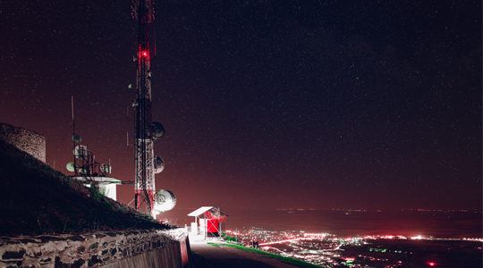 A telecommunication tower stands illuminated at night on a hilltop, with a backdrop of a starry sky and sprawling city lights in the distance.