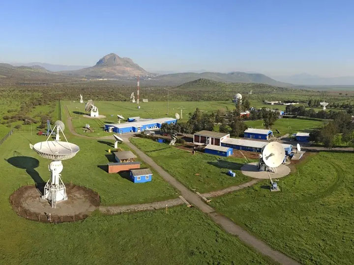 A satellite station with lush greenery, featuring a series of large dish antennas and smaller structures, under a clear blue sky with a mountainous backdrop.
