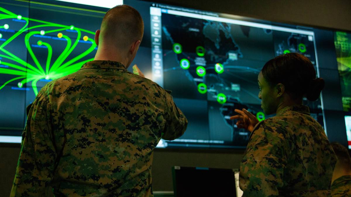 Two military personnel are examining a large digital screen displaying a world map with various data points and graphics.