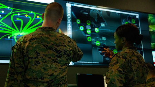 Two military personnel are examining a large digital screen displaying a world map with various data points and graphics.