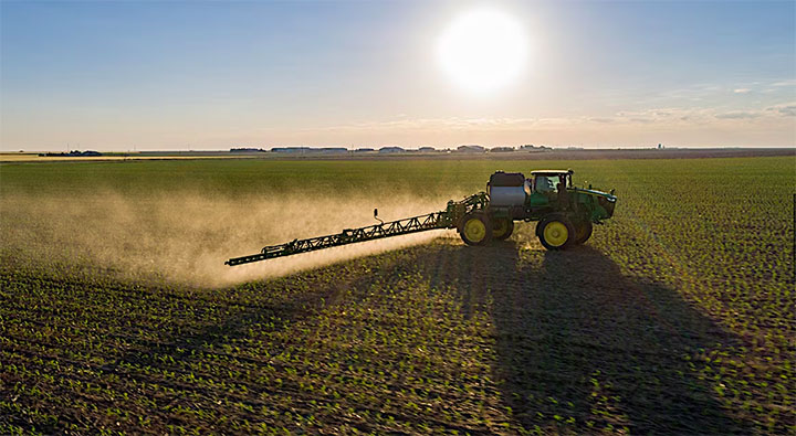 A tractor equipped with a long boom sprayer operates in a vast field, distributing agricultural chemicals during a bright, sunny day.