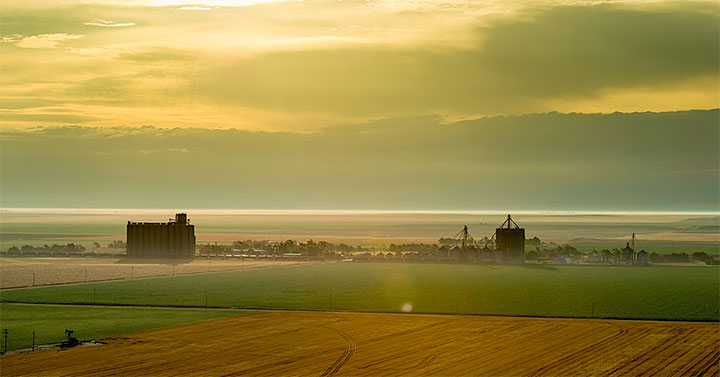 Sunrise over a rural landscape featuring expansive farmland with varying shades of green and gold crops, large silos, and farm buildings in the distance under a hazy sky.
