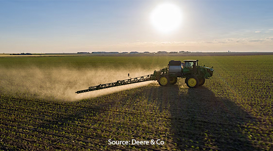 A tractor equipped with a long boom sprayer operates in a vast field, distributing agricultural chemicals during a bright, sunny day.