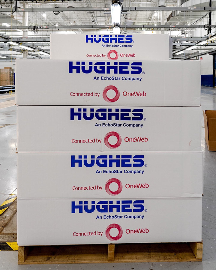 A stack of boxes with the "Hughes An EchoStar Company" and "Connected by OneWeb" logos, placed on a wooden pallet in a warehouse.