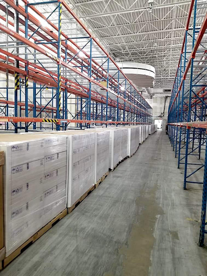 A warehouse interior with rows of tall, empty blue and orange shelving on the right, and a long line of wrapped pallets containing Hughes OneWeb antennas on the left, ready for shipment.