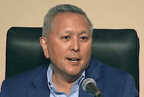 A man with short gray hair, wearing a blue shirt and speaking into a microphone, appears engaged in a discussion at an event.