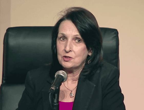 A woman with medium-length dark hair, wearing a black blazer over a pink blouse, speaking into a microphone at a panel discussion.