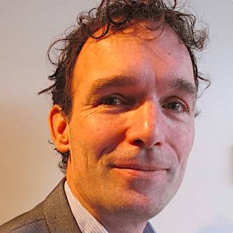 A close-up portrait of a smiling man with curly dark hair, wearing a gray suit jacket, against an indistinct background.