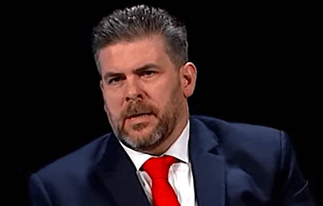 A man with short grey hair, wearing a dark suit and red tie, against a black background.