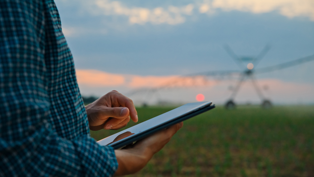 Hands holding a large tablet in a field with irrigation equipment in the background.