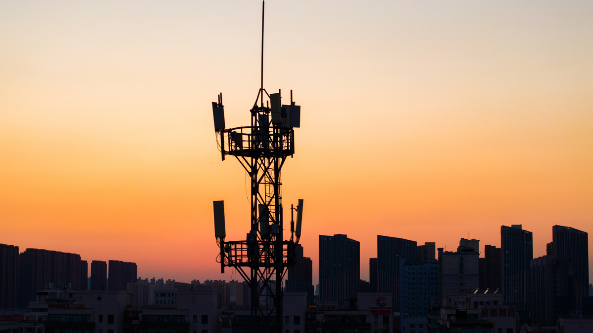 A telecommunications tower silhouetted against an orange and yellow sunset, with a dark city skyline in the background.