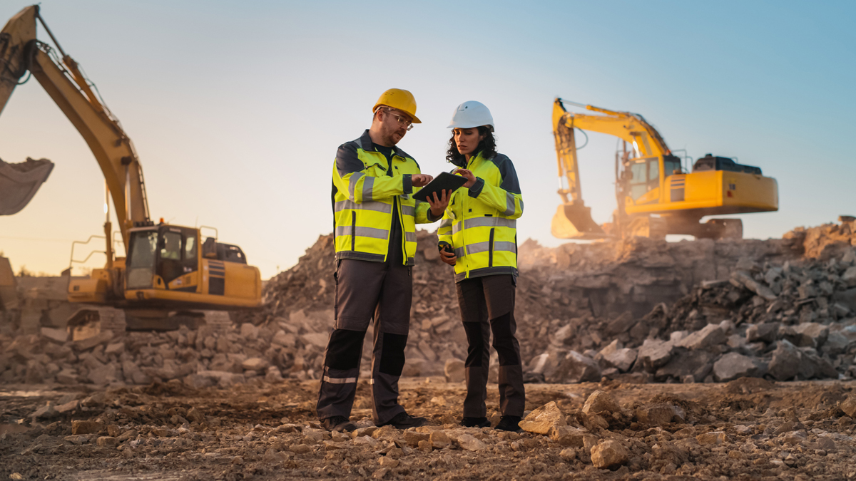 Two construction workers in safety vests and hard hats consult a tablet on a worksite with two yellow excavators in the background.
