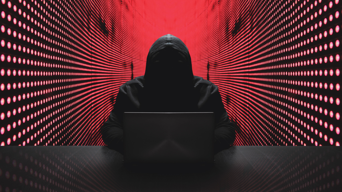 Anonymous hooded figure using a laptop, representing a hacker, against a red digital background of radiating data lines.