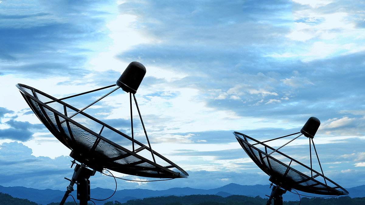 Two large satellite dishes are silhouetted against a cloudy sky with mountains in the background, representing communication and data transmission.