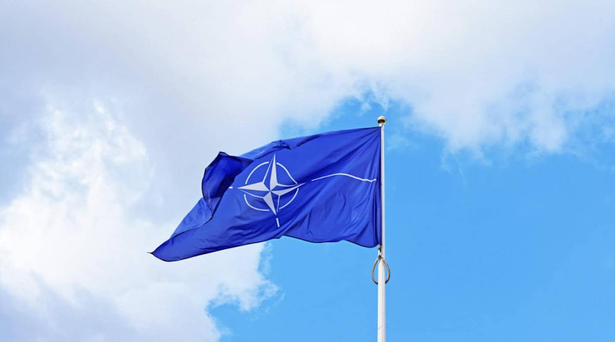 The flag of the North Atlantic Treaty Organization (NATO) waving against a bright, partly cloudy sky.