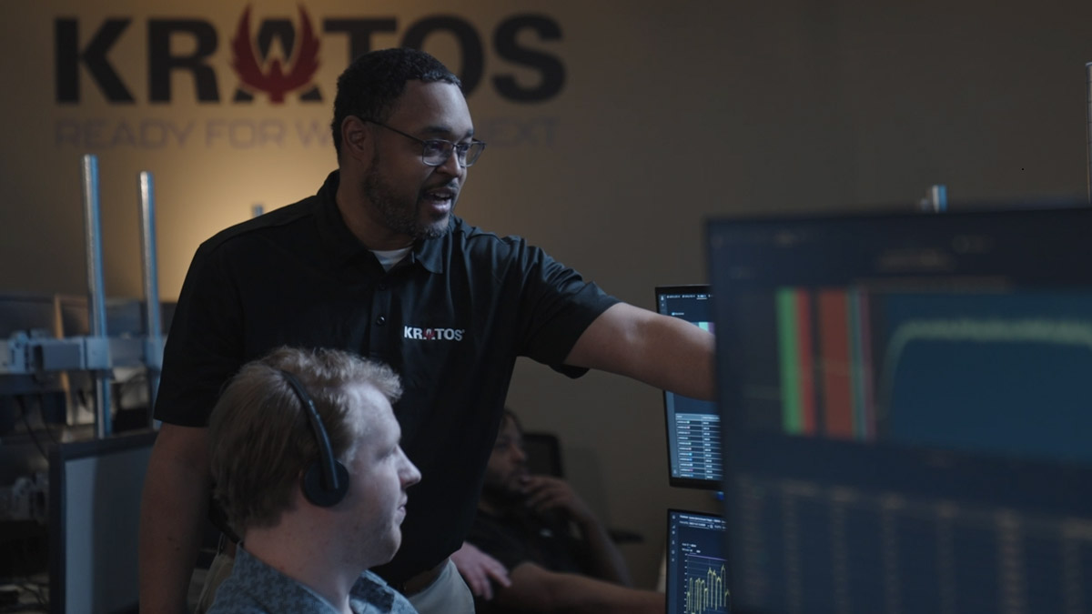 A man in a Kratos shirt points to a computer monitor while assisting a colleague wearing a headset in a control room, with multiple screens displaying data charts and graphs.